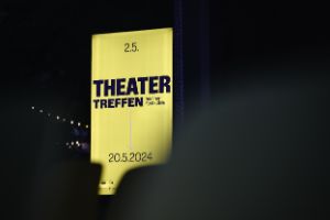 Ein beleuchtetes gelbes Poster mit dem Schriftzug „Theatertreffen“ sowie eine Lichterkette und Silhouetten von Menschen in einer dunklen Umgebung. 