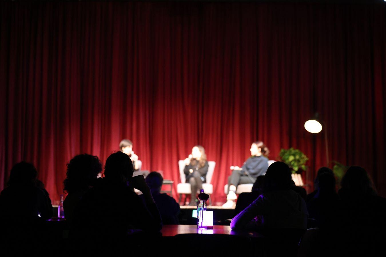 Hinter den Schatten des Publikums sieht man verschwommen drei Personen auf der Bühne vor einem roten Vorhang. Sie sitzen auf Stühlen und halten Mikrofone.