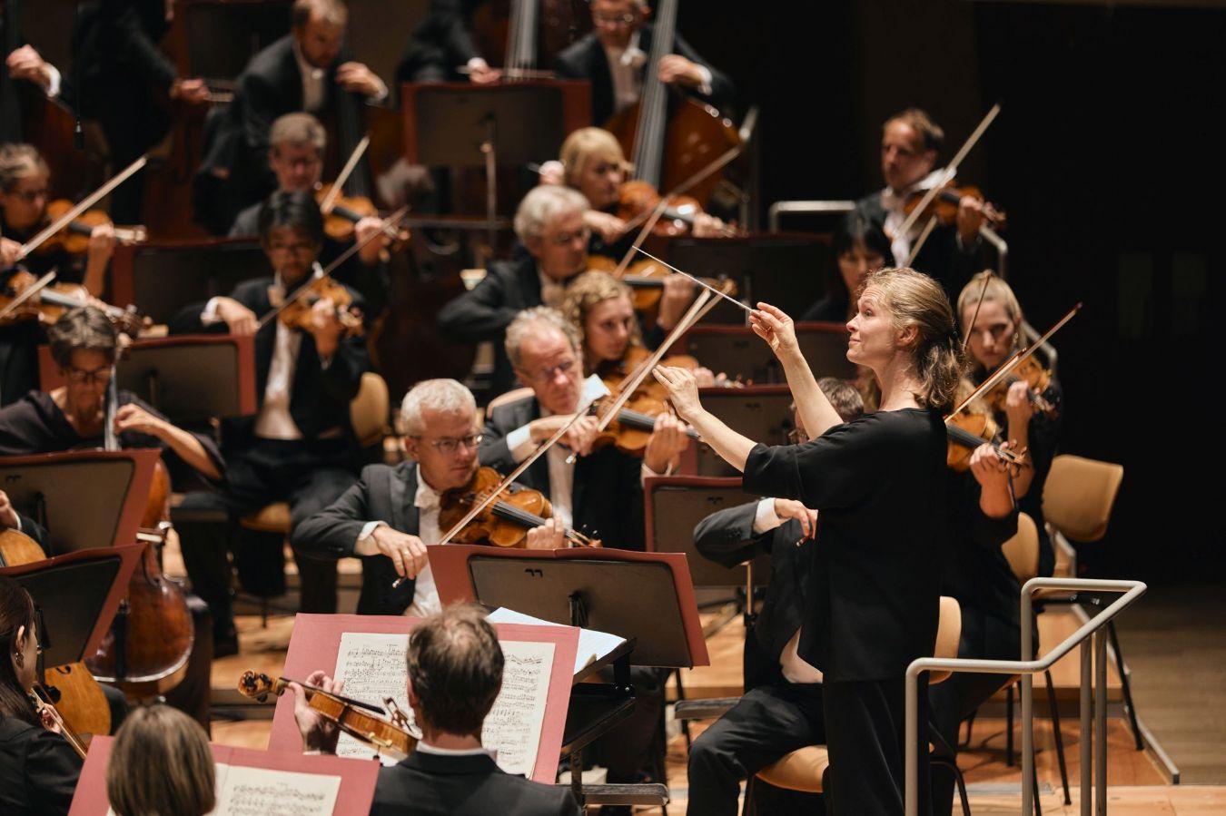 Mehrere Personen machen gemeinsam Musik. Die meisten von ihnen spielen eine Geige. In der Mitte steht ein*e Digirent*in, er*sie leitet das Orchester mithilfe von Handzeichen.