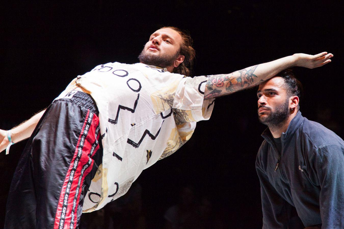 Zwei Jungen tanzen gemeinsam. Der linke Junge beugt sich nach hinten und streckt dabei seinen Arm über den Kopf des andere Jungen aus. Sie tragen beide lockere Kleidung.