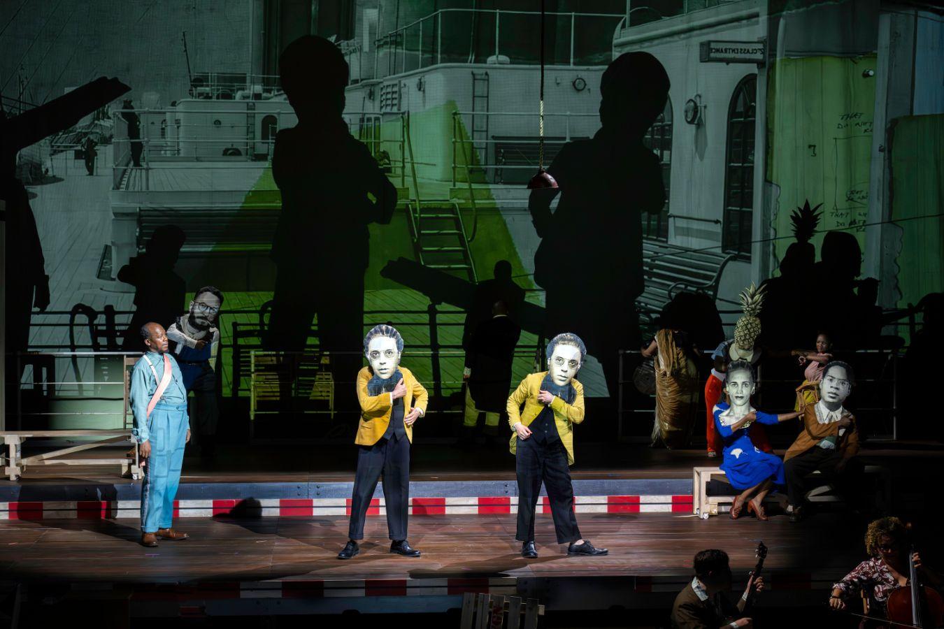 Personen mit Masken, die Porträts von historischen Personen abbilden. Dahinter eine Projektion eines Schiffsdecks.