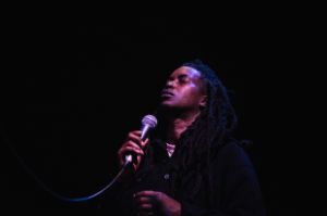 Camae Ayewa aka Moor Mother during a live performance. The stage is dark, the spoken word artist's face is softly lit. She holds a microphone in her right hand. Ayewa's eyes are closed, her face tilted slightly upwards.
