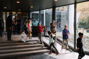Children run down the ramp to the ticket hall of the Haus der Berliner Festspiele. A boy sits on the top step. Next to him stands a man dressed in black. Some children run up the stairs, holding on to the banisters.