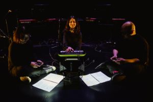 Three people sit on a stage. Elif Kara sits in the centre at a toy piano, with musicians Bertram Wee and Lynette Yeo sitting to her right and left with controllers in their hands.