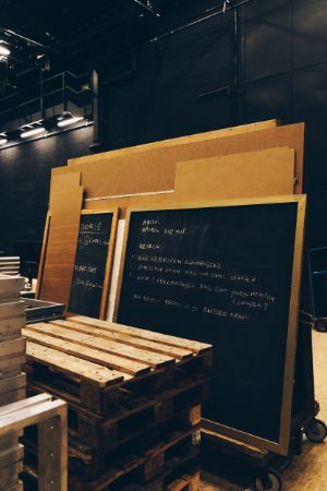 In a room there are Euro pallets, plywood boards and panels in front of a black wall. On the two front boards, among other things, rules have been written down in chalk.