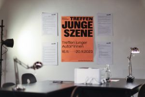 An orange poster with the inscription “Treffen junge Szene” hangs on the wall behind two desks.