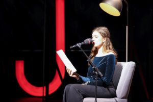 Lilli Nawar sits in an armchair and speaks into the microphone in front of her. In the background is a reading lamp and a glowing red “J”.