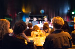 Young people sitting at tables, watch a conversation on a stage.