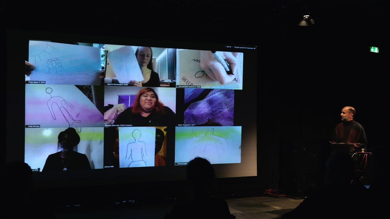 Projection of a video call screen on a stage. Not all people can be seen. Most of them are holding drawings of human bodies to the camera. A moderator is at the edge of the stage, and several audience members can be seen in the foreground.