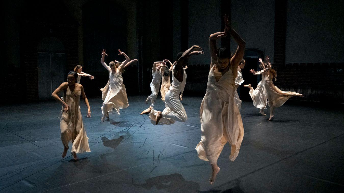 Auf einer leeren Bühne springt eine Gruppe von Tänzerinnen in weißen Kleidern in die Höhe.