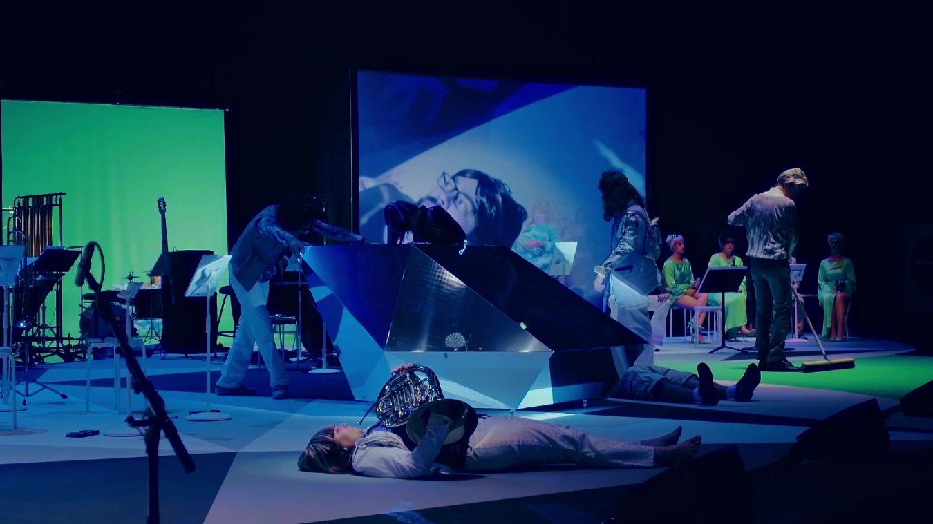A stage with various musical instruments and video projections. In the foreground, a person with a horn lies on the floor.