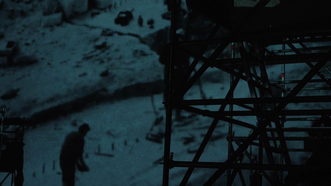 Ein Baugerüst steht vor einer Leinwand, die das Fundament eines Hauses zeigt, auf dem Bauarbeiter herumlaufen