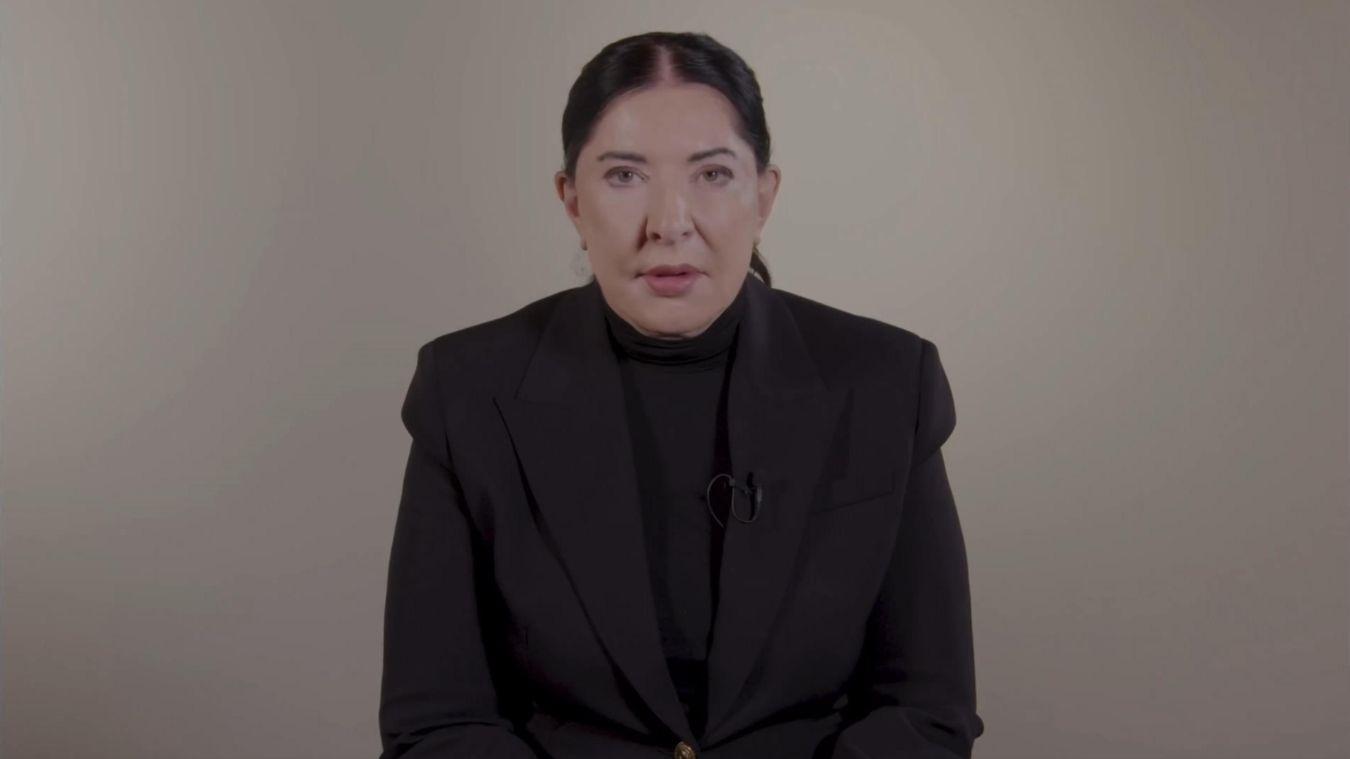 A person is seated in front of a neutral beige background. They are wearing a dark blazer over a dark top, with a clip-on microphone attached to the lapel. The person is sitting calmly, facing the camera.
