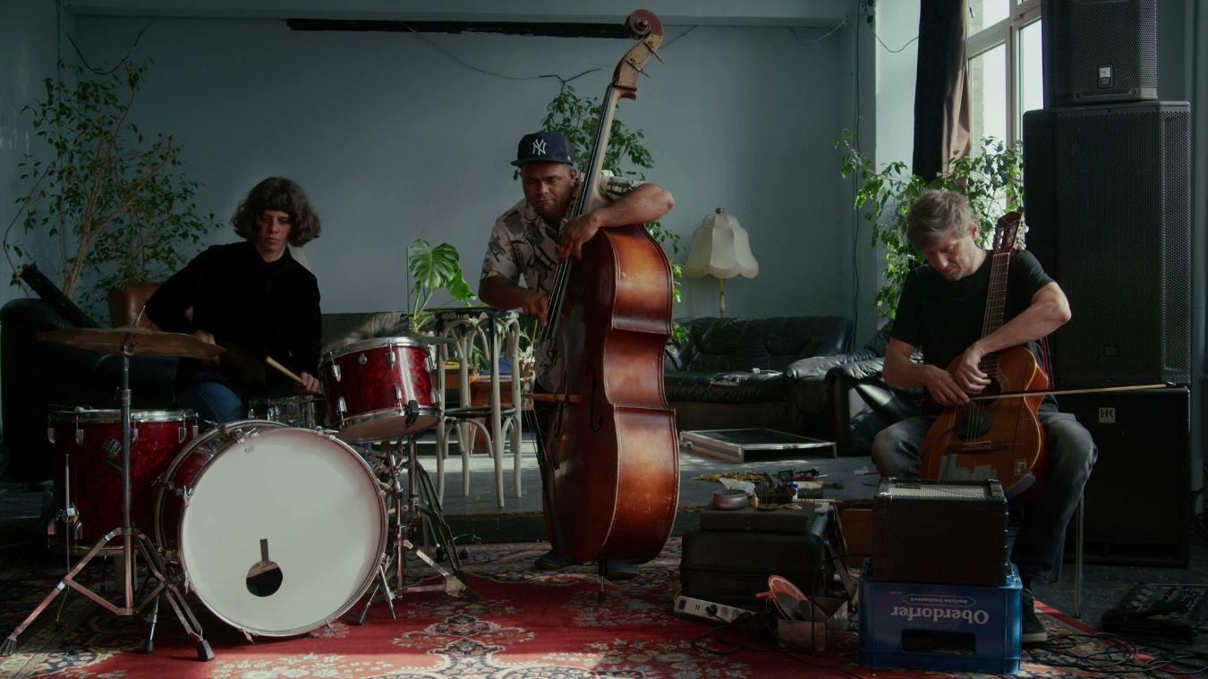 Three musicians with drum kit, double bass and cello in a room with plants, carpet and speakers