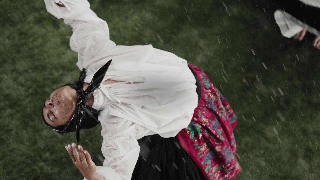 A person dances on grass in traditional clothing, arms extended in a dynamic pose.