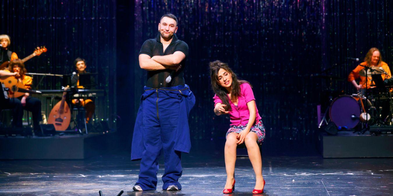 Ein Mann steht mit verschränkten Armen neben einer sitzenden Frau im pinken Kostüm, dahinter Menschen mit Instrumenten.