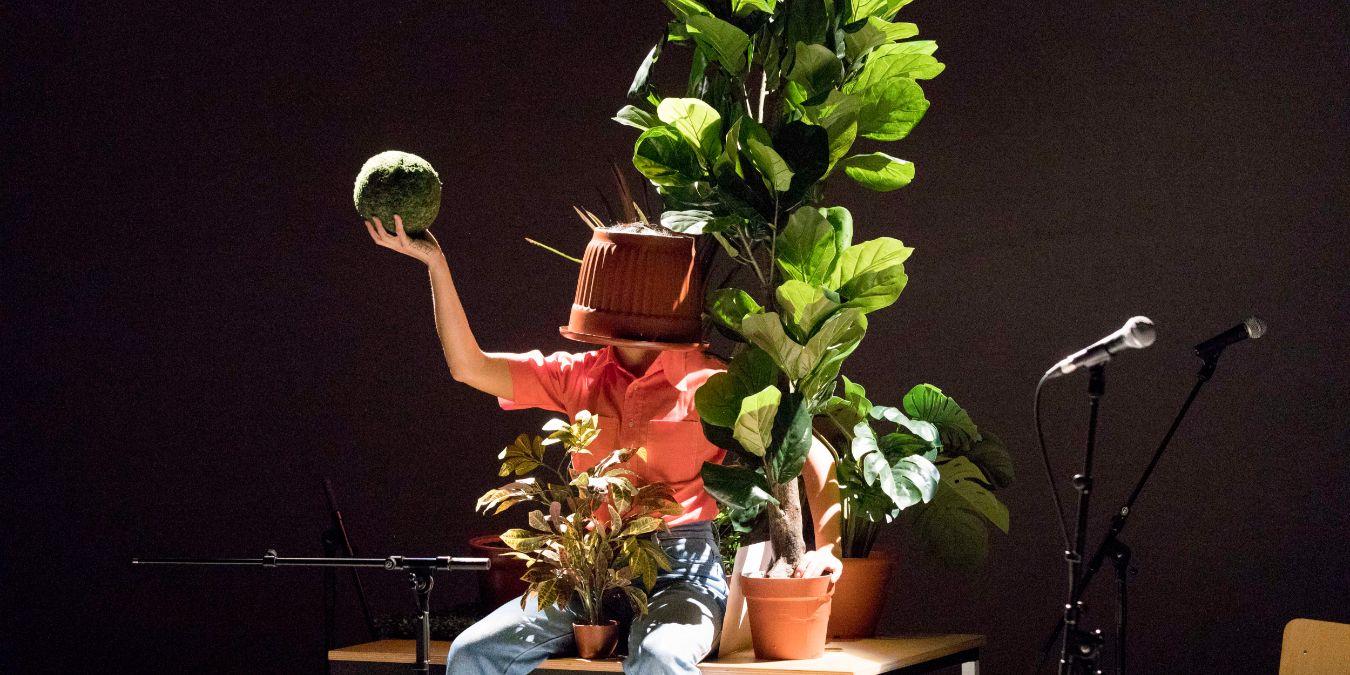 Eine Frau mit einem Blumentopf auf dem Kopf ist von Pflanzen umgeben.