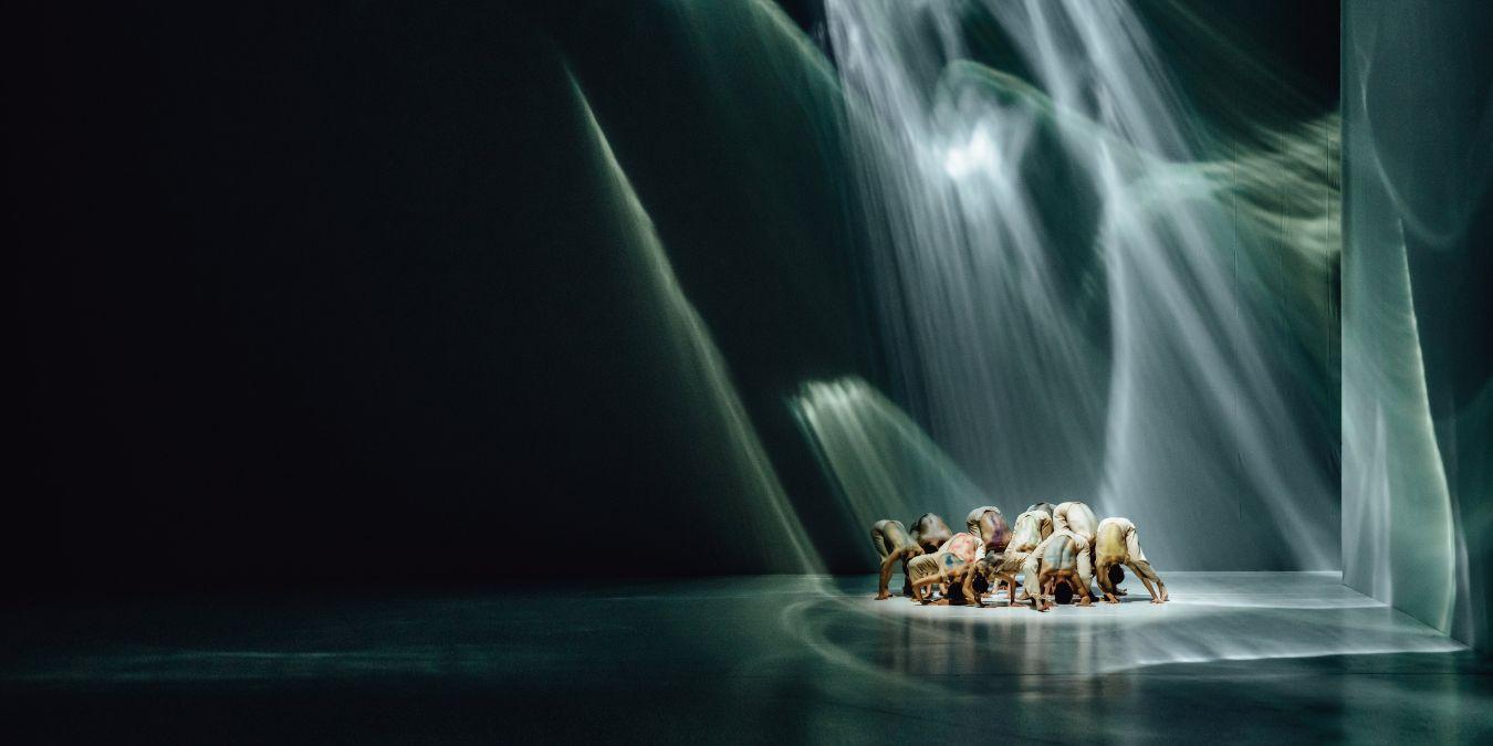 Mehrere Personen kauern dicht beieinander auf einer Bühne unter dramatischen Lichtstrahlen.