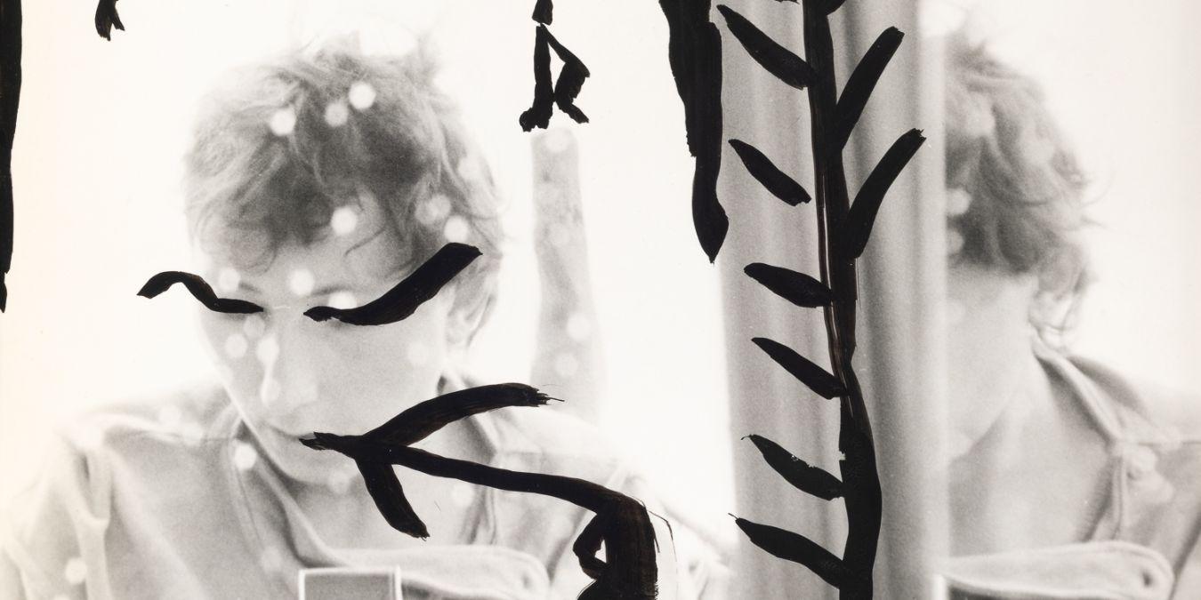 Ein mit schwarzen Mustern bemaltes Selbstporträt von Gabriele Stötzer, die sich mit einer Analogkamera im Spiegel fotografiert.