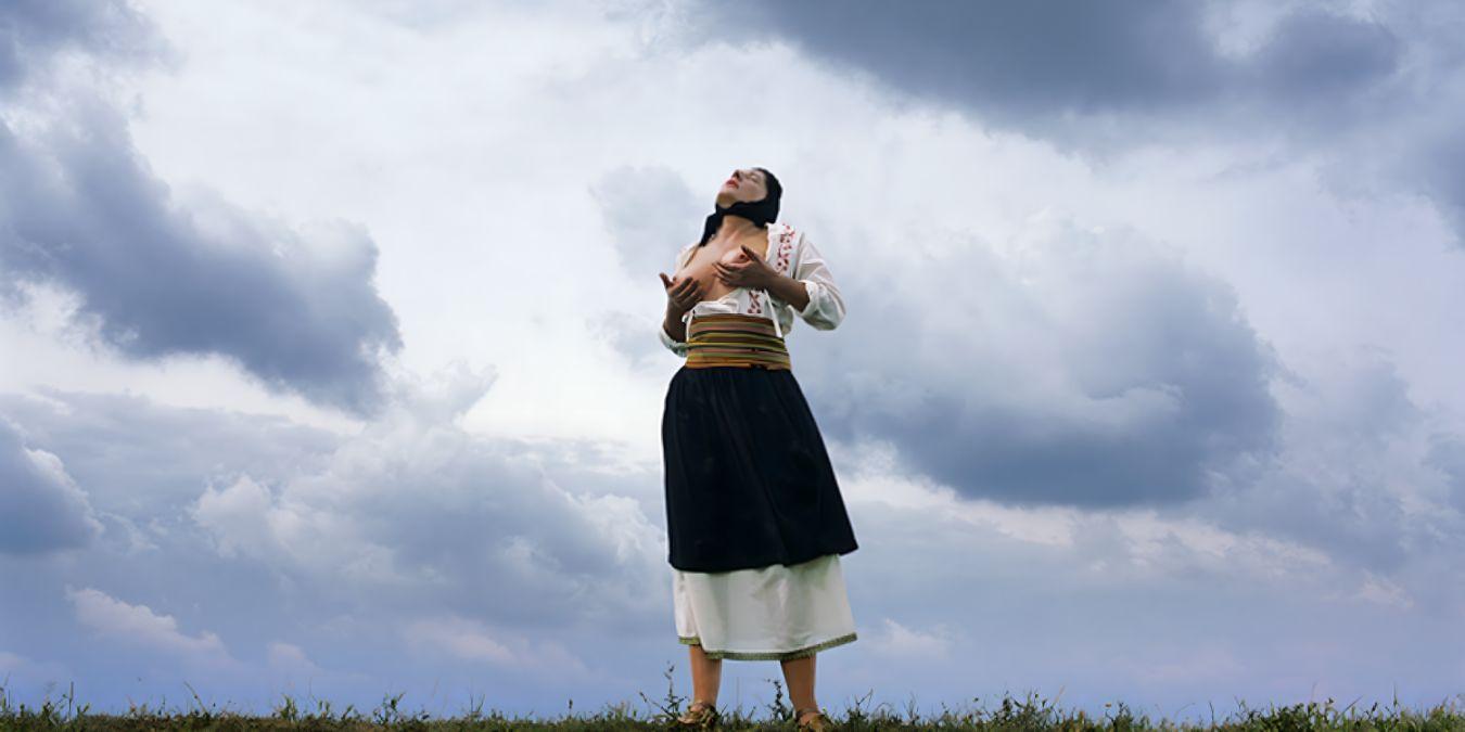 Eine Person mit einem schwarzen Tuch auf dem Kopf massiert sich im Freien ihre nackten Brüste.