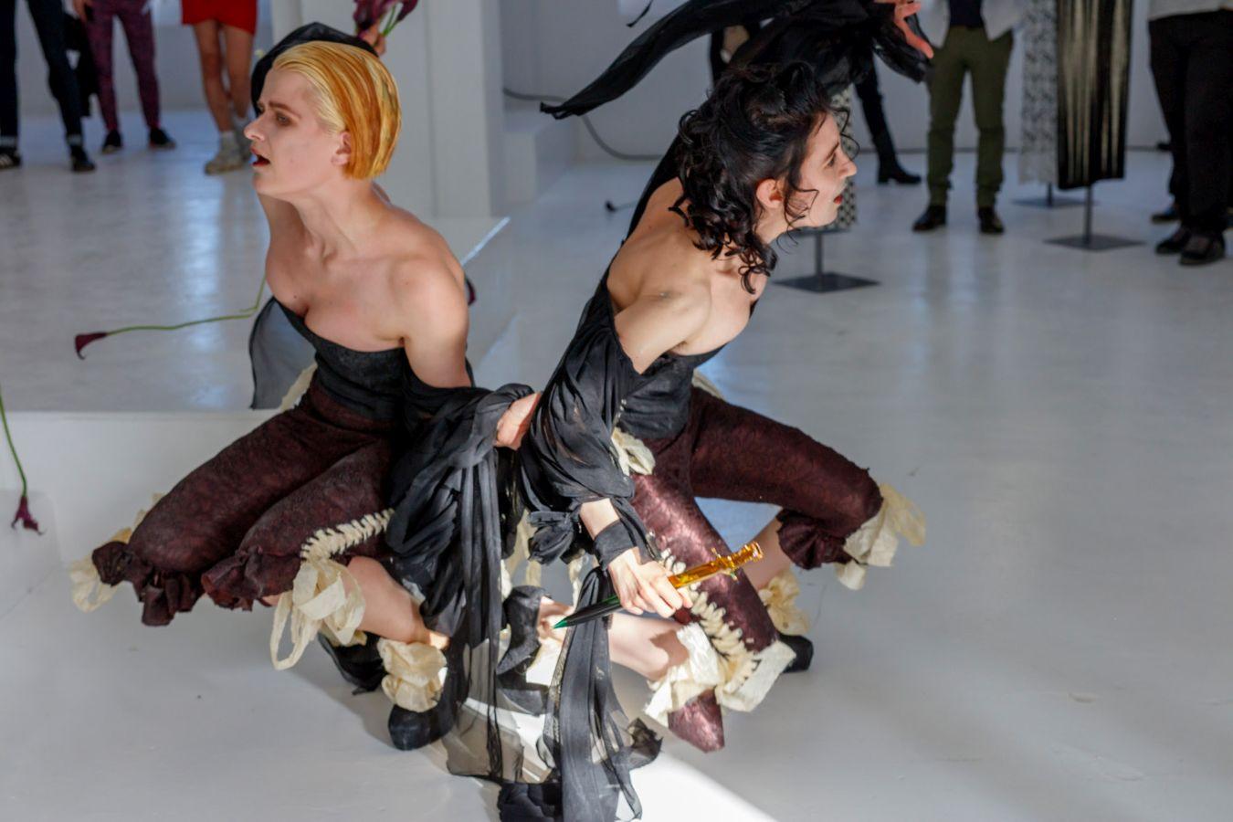 Two people during a performance, both squatting back to back.