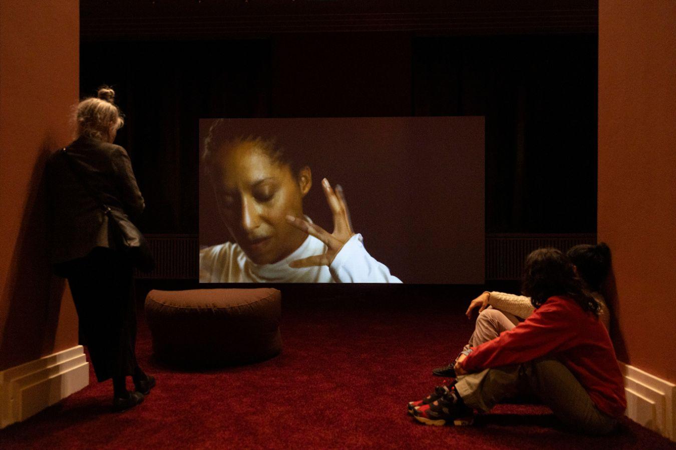 In a dimly lit room with dark red velvet carpet, a large screen displays Ligia Lewis’s A Plot, A Scandal. Two visitors face the screen, where the faint outline of a person in a white dress is visible