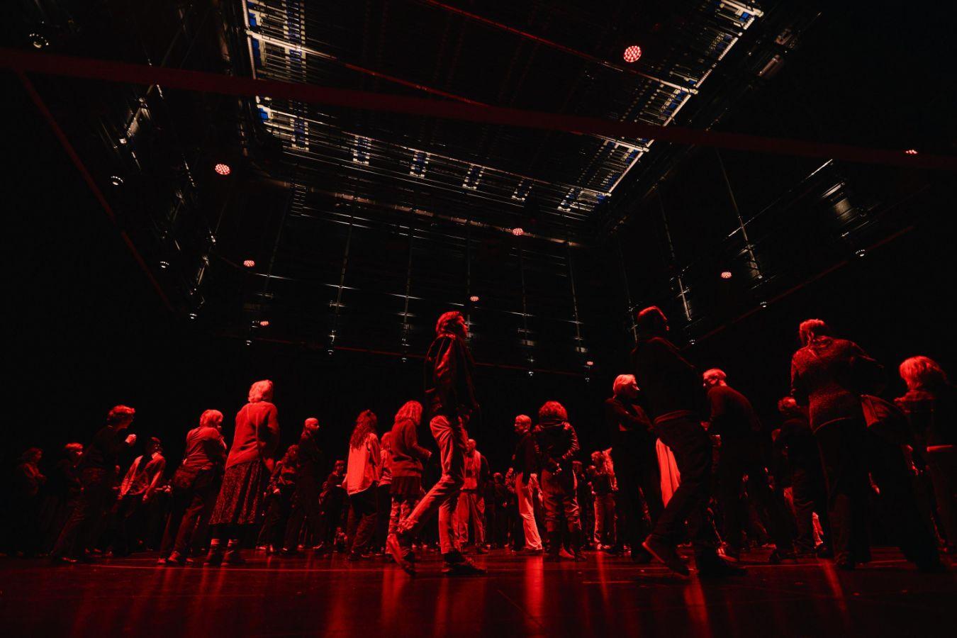 The audience moves around the stage. The flyloft is above them.