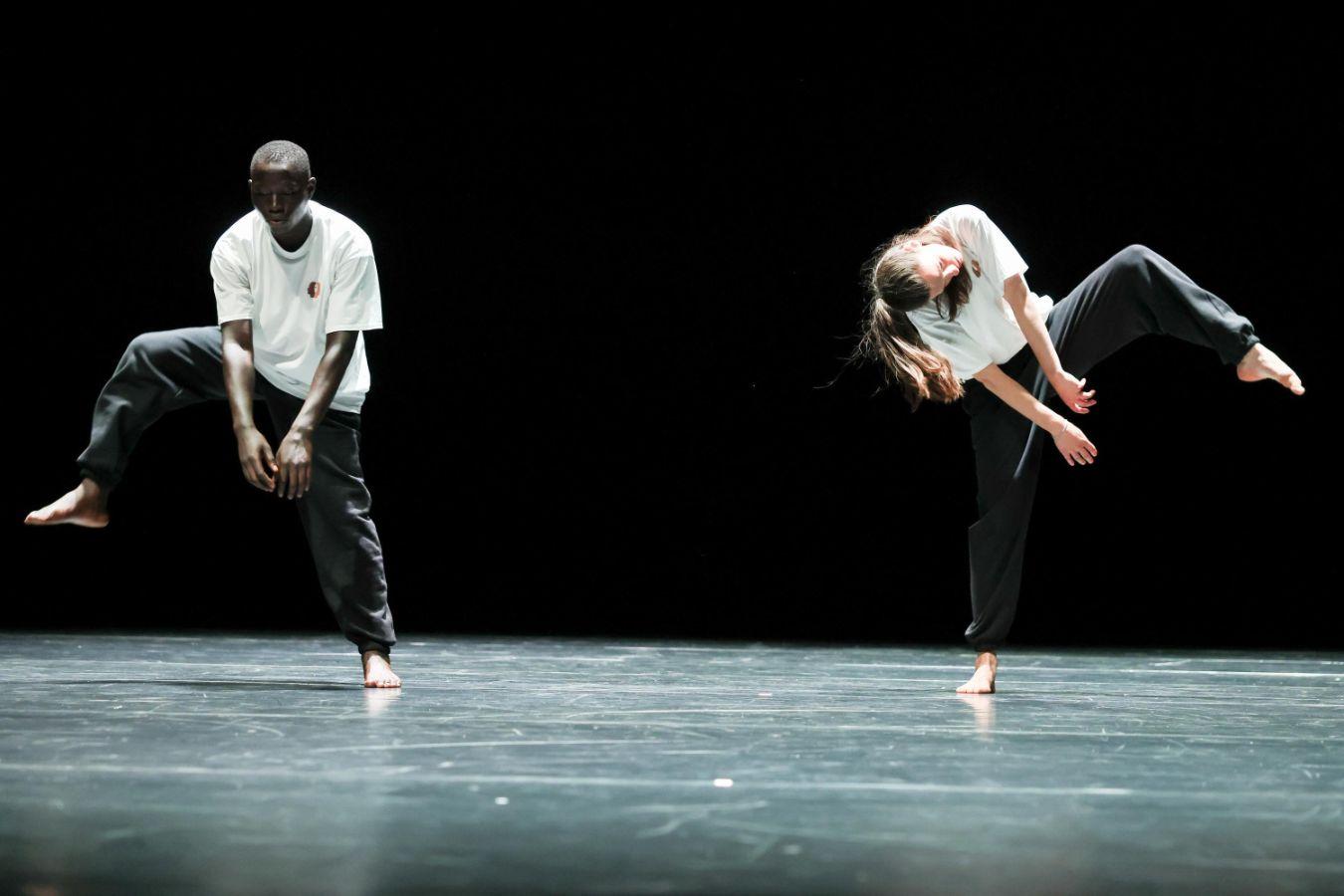 Two dancers stand apart on a large stage with a dark floor and background. Both lift one bent leg and extend their arms downward.