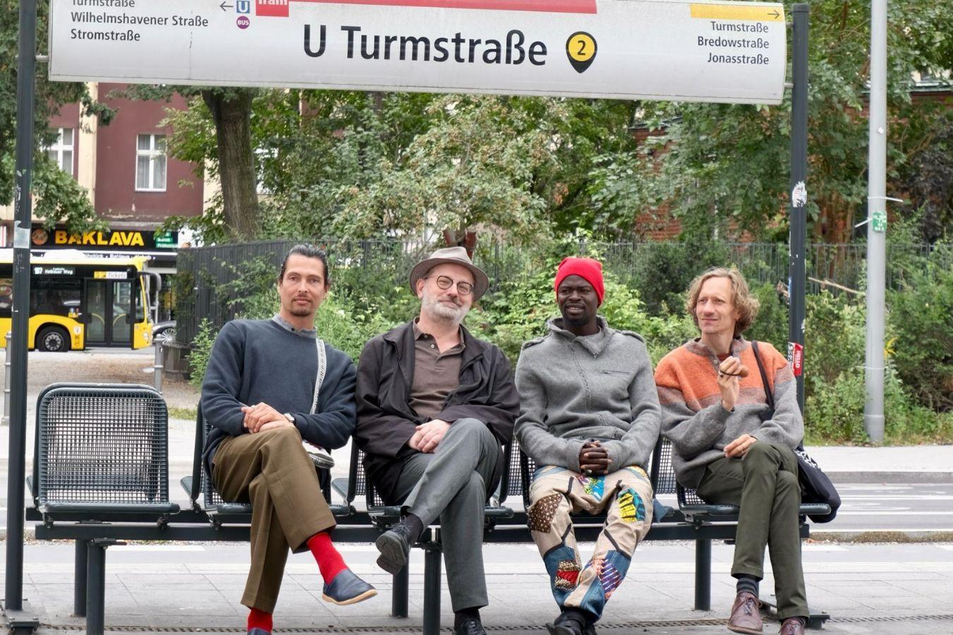 Simon Sieger, Michael Griener, Assane Seck, Joel Grip sit on a bench, above them the sign for the U-Bahn station "U Turmstraße"