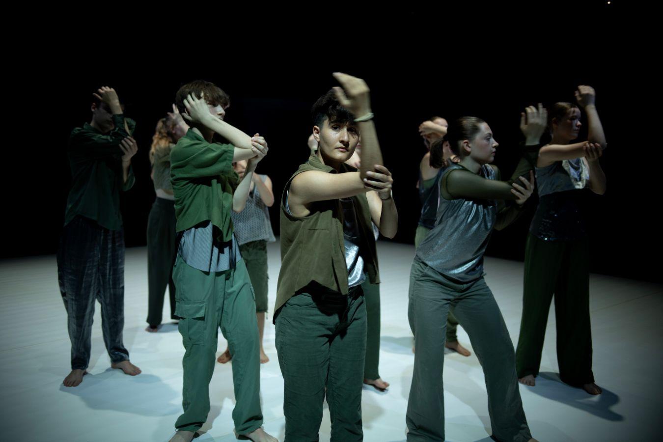 Several dancers stand in rows on the stage, all with their right arm raised and bent. The person in the front row looks at the camera, the others look to the side.