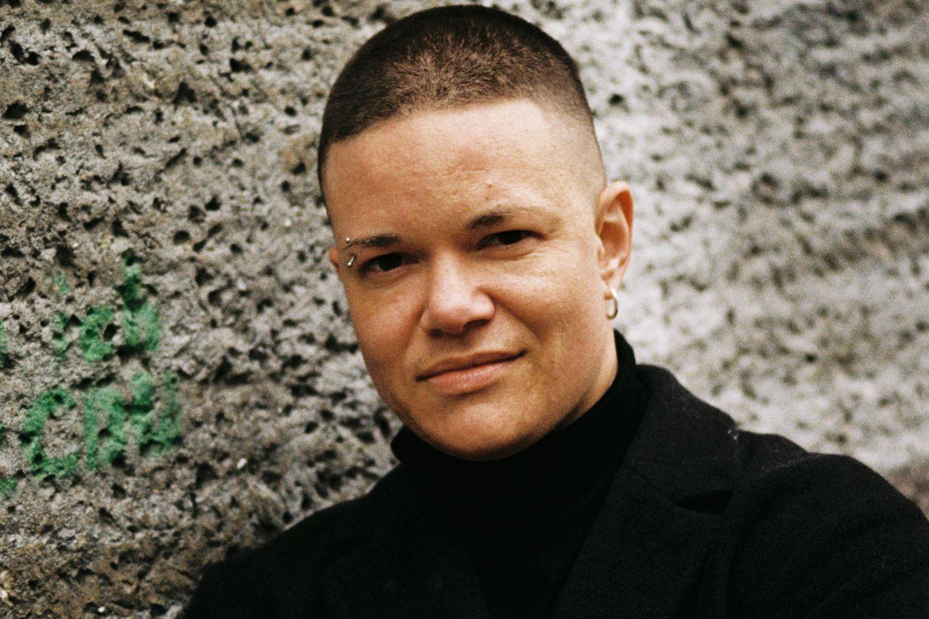 Porträtaufnahme von einer Person mit verschränkten Armen vor einer Steinwand