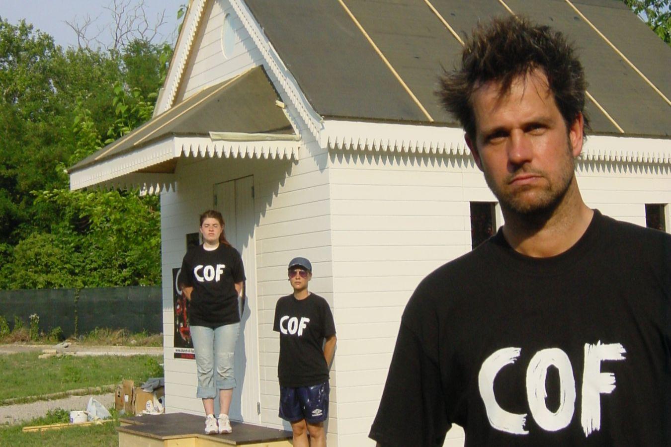 Christoph Schlingensief is standing in front of a white wooden chapel. Behind him are two people, all of them wearing T-shirts with the letters “COF” printed on them.