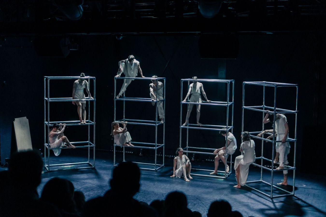 Four silver scaffolds stand next to each other on a dark stage, on which several dancers sit or stand. They wear white, simple costumes.