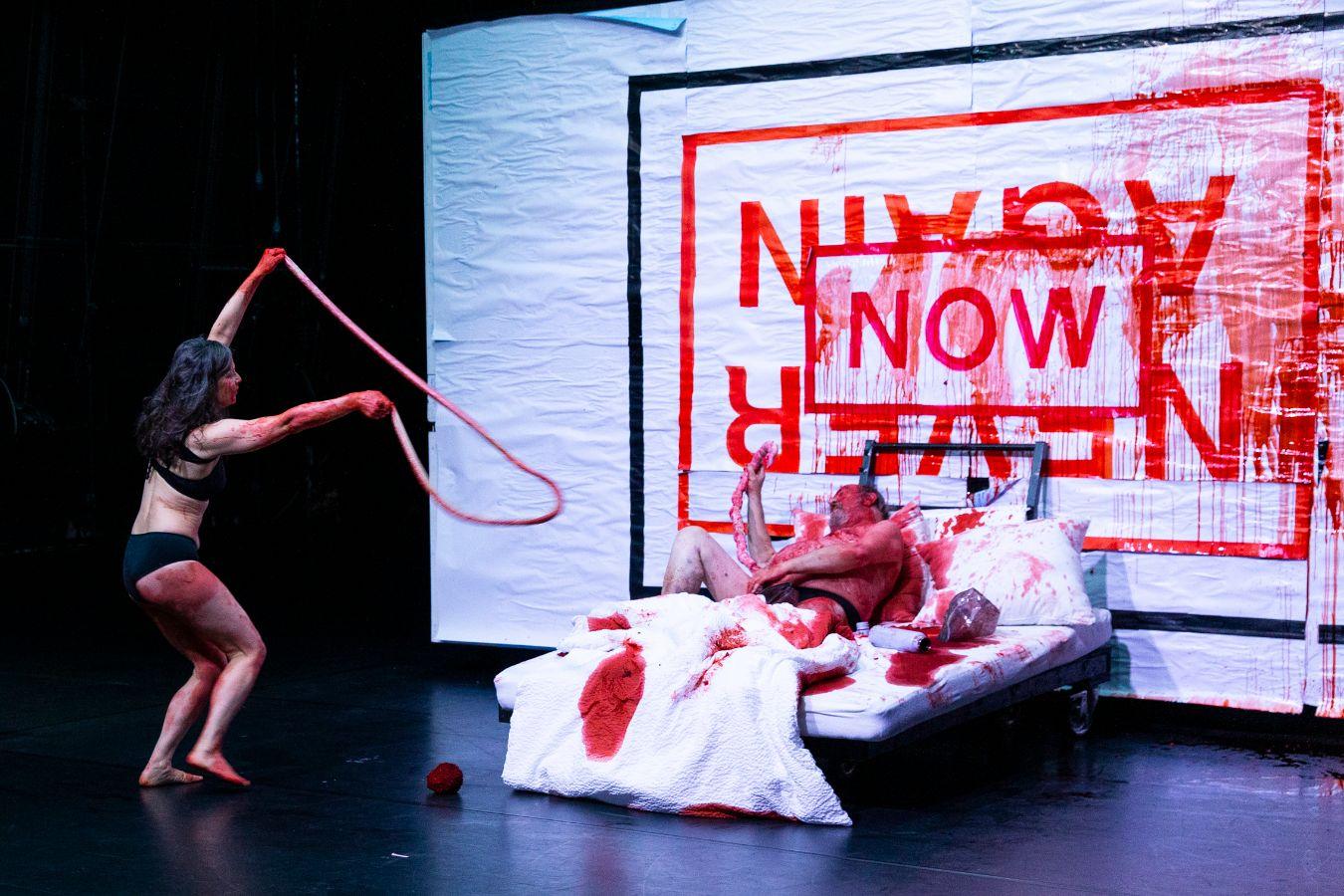 Tanzende Frau in Unterwäsche mit Gedärmen und blutverschmierter Mann im Bett vor einer Plakatierung mit der Aufschrift "Now" und "Never Again".
