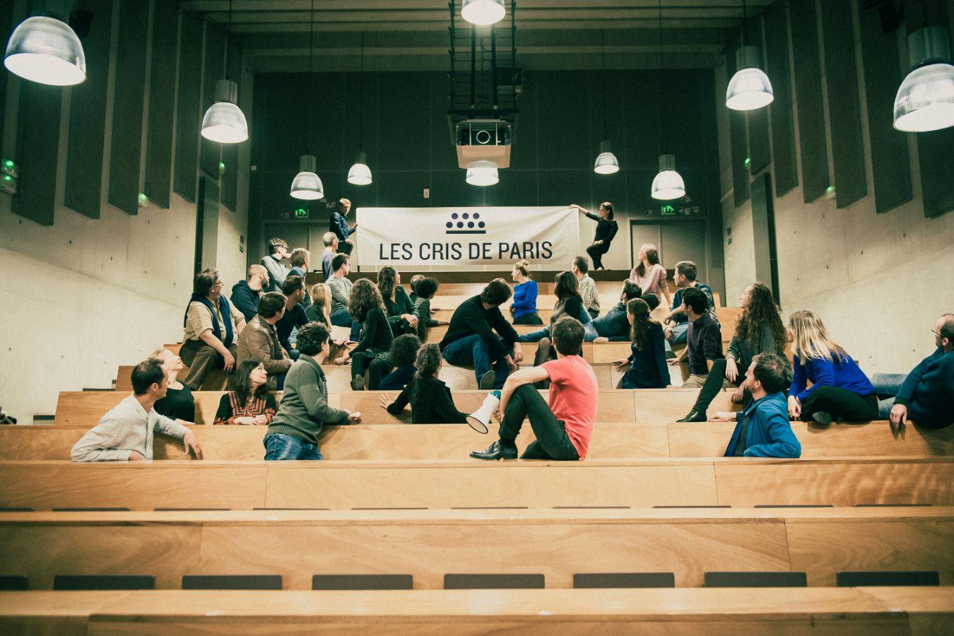 Oben auf einer Tribüne halten zwei Personen ein Banner mit der Aufschrift "LES CRIS DE PARIS", das von vielen Menschen in den unteren Reihen betrachtet wird.