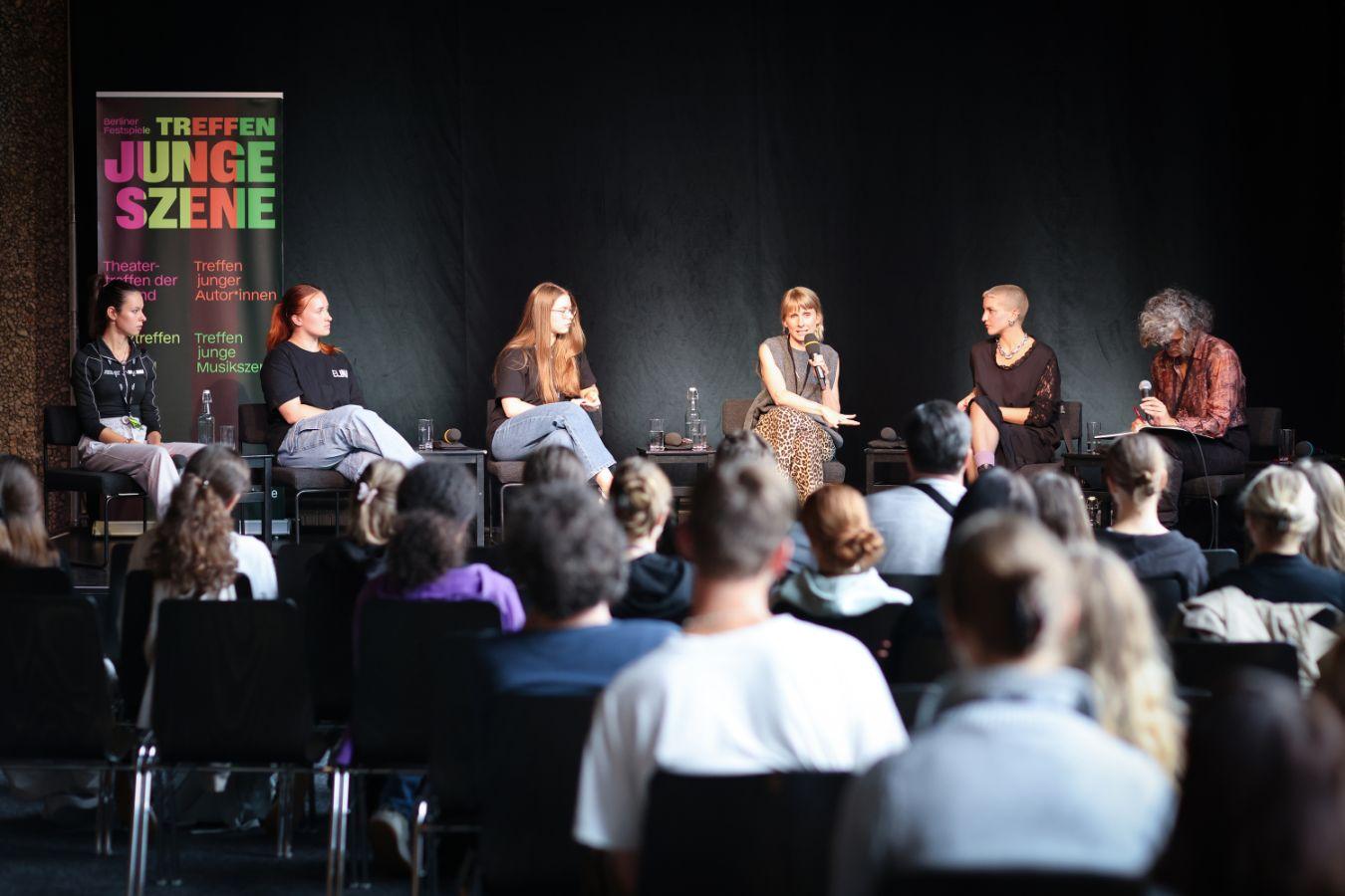 Six people sit on stage, the audience sits on the opposite side