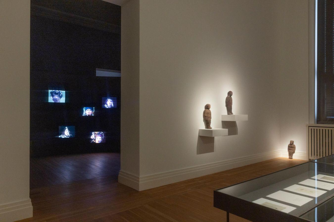 In einem Ausstellungsraum stehen kleine Figuren an der Wand und rechts ist eine Glasvitrine angeschnitten. Durch einen Durchgang ist der nächste Ausstellungsraum zu sehen.