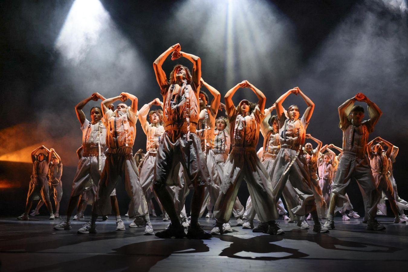 A group of dancers stands on a dark stage in a powerful pose. From above, spotlights cast bright light onto them.