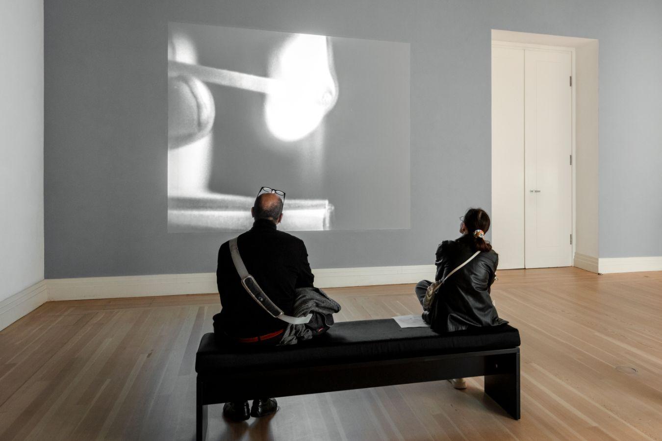A video of a person with a match is projected onto a wall in an exhibition space, with two people sitting on a bench in front of it.