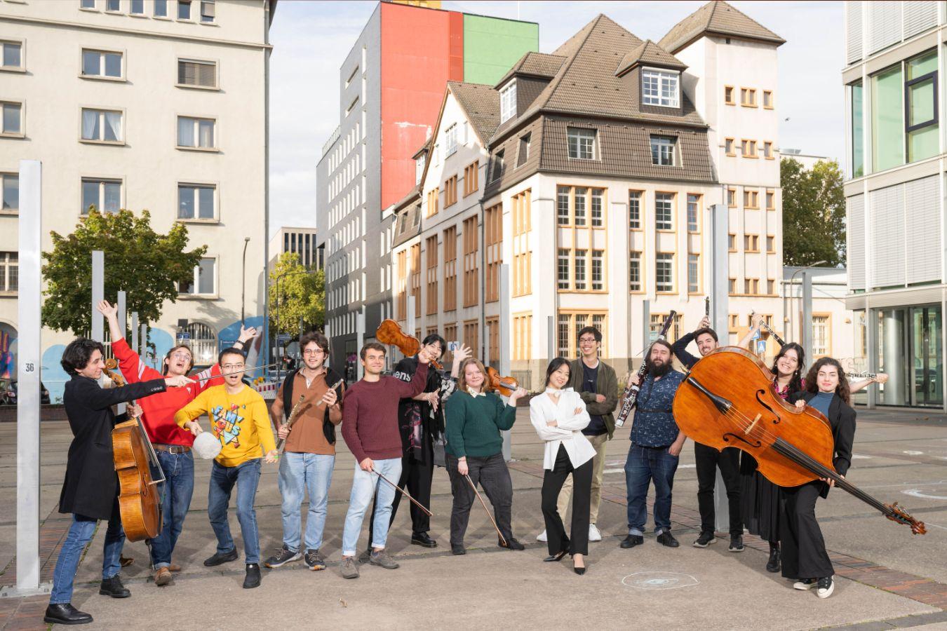 13 Musiker*innen bewegen sich mit ihren Instrumenten auf einem städtischen Platz