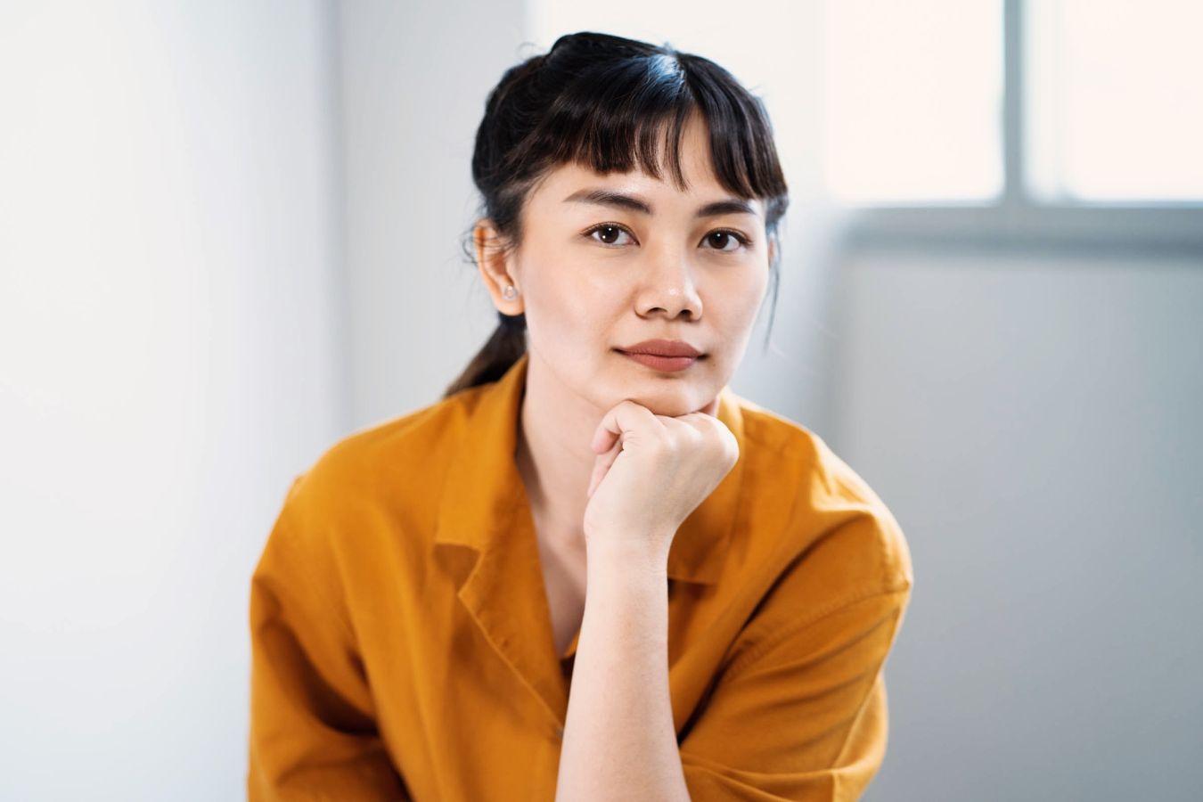 A woman with dark brown hair in a ponytail and a yellow blouse resting her face on her fist