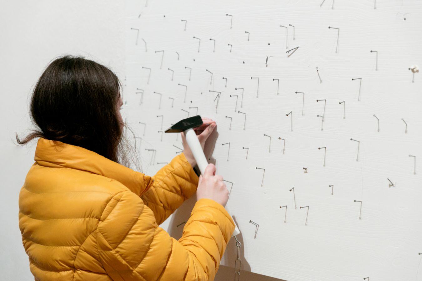 A person uses a hammer to hammer nails into a white canvas in which some nails are already stuck.