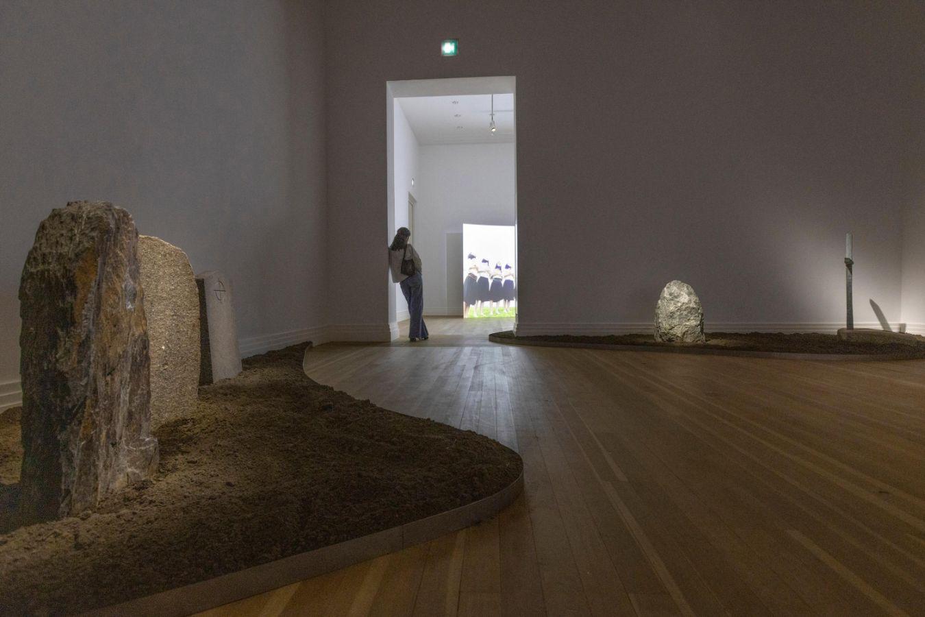 In einem Ausstellungsraum stehen links und hinten rechts Grabsteine. Im Hintergrund steht eine Person im Durchgang zum nächsten Raum.