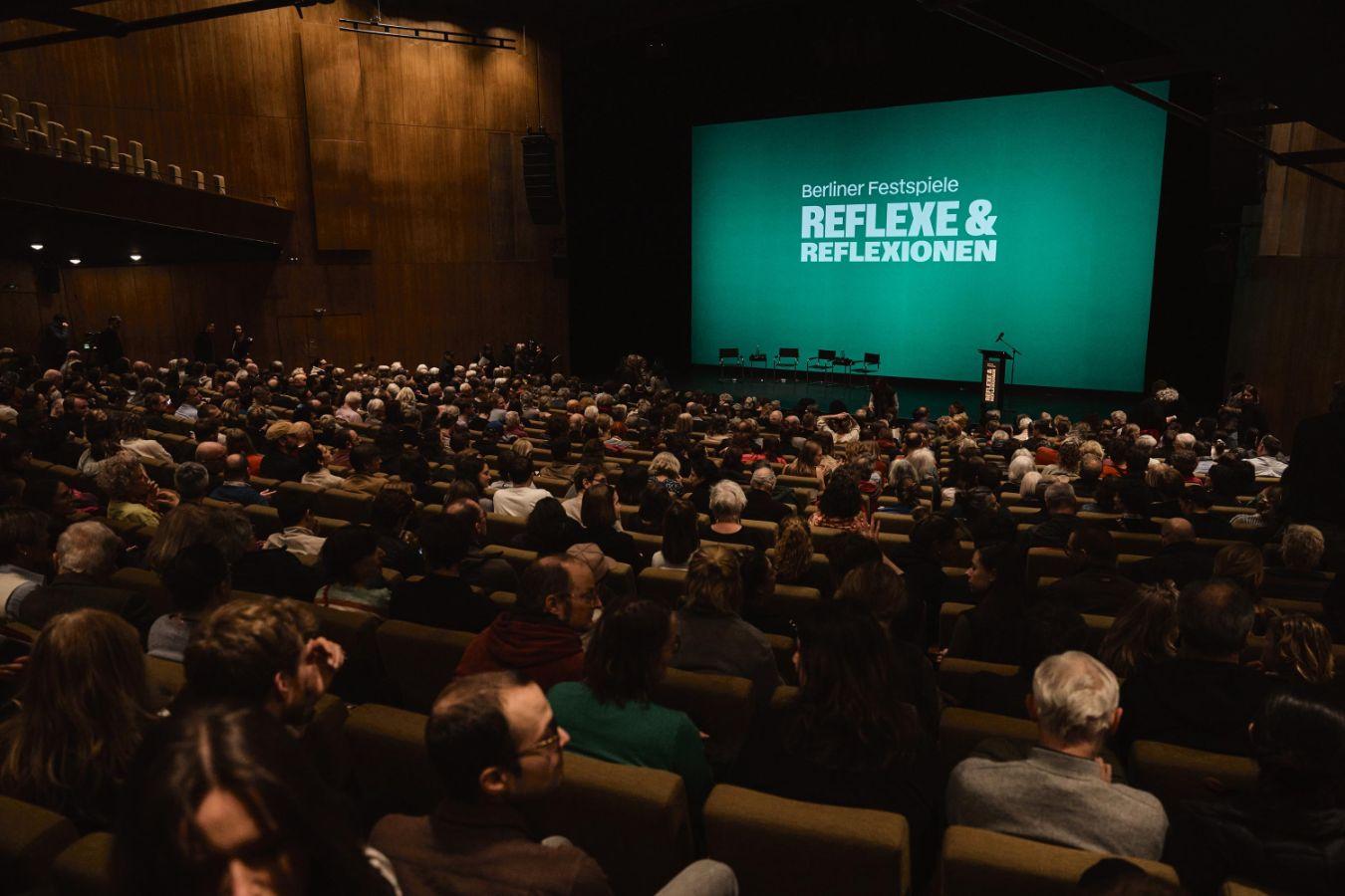 Publikum sitzt in einem großen Theatersaal und blickt auf eine Bühne mit grünem Bildschirm mit der Aufschrift „Berliner Festspiele – Reflexe & Reflexionen“.