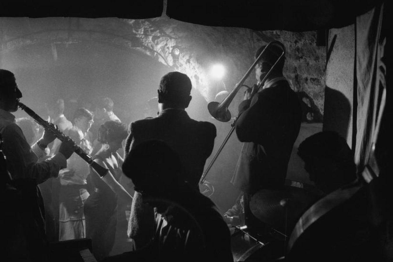 Drei Männer spielen auf Blasinstrumenten in einem Kellergewölbe. Sie sind von hinten zu sehen, vor ihnen stehen Menschen.