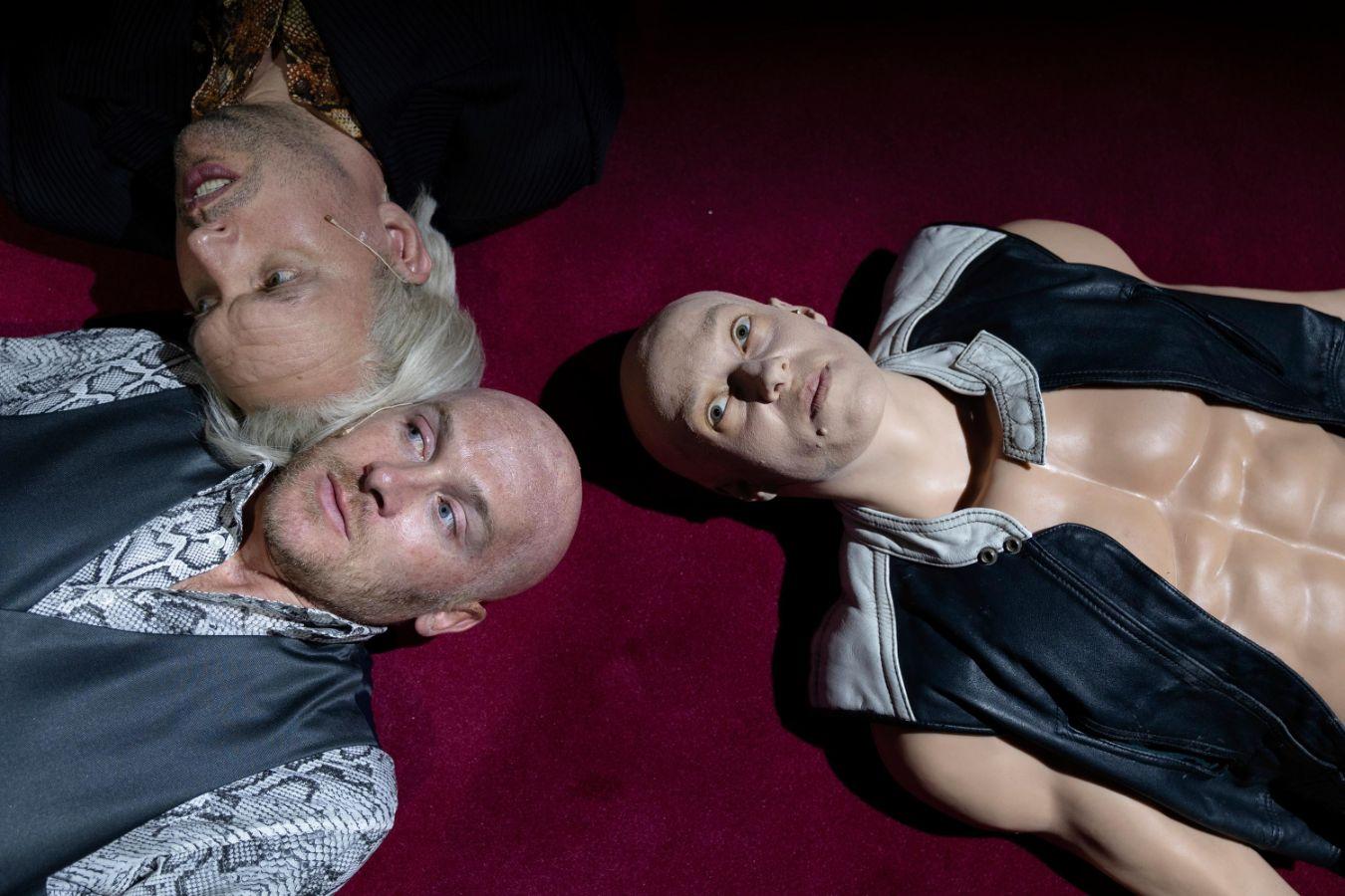 Three figures lie side by side on a dark red surface, facing the camera.