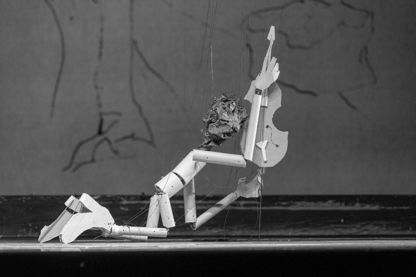 Eine Marionette hält, auf den Knien abgestützt, ein Streichinstrument in die Höhe.