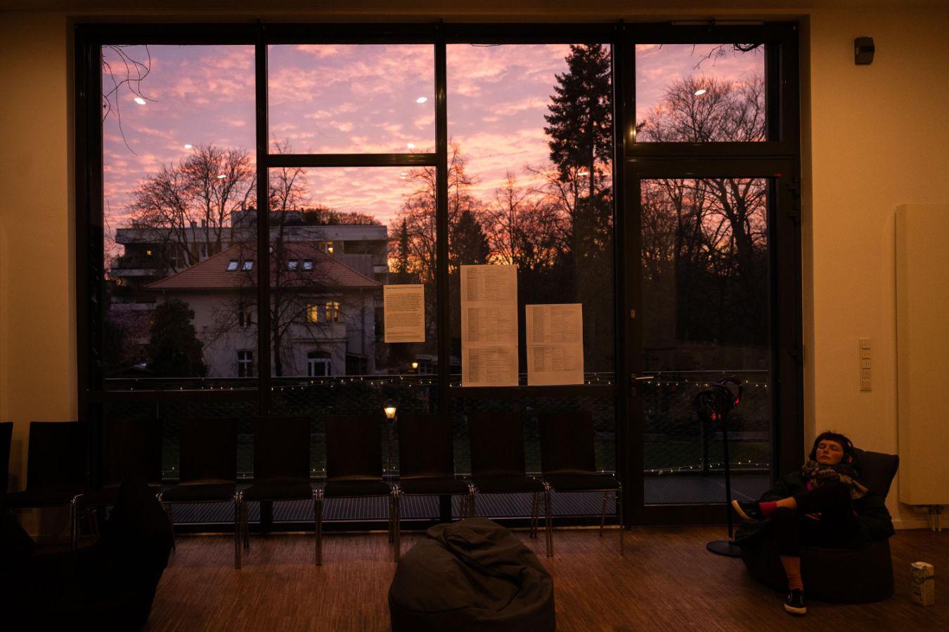 Eine Person sitzt in einem Sessel vor einer Fensterfront in der Abenddämmerung.
