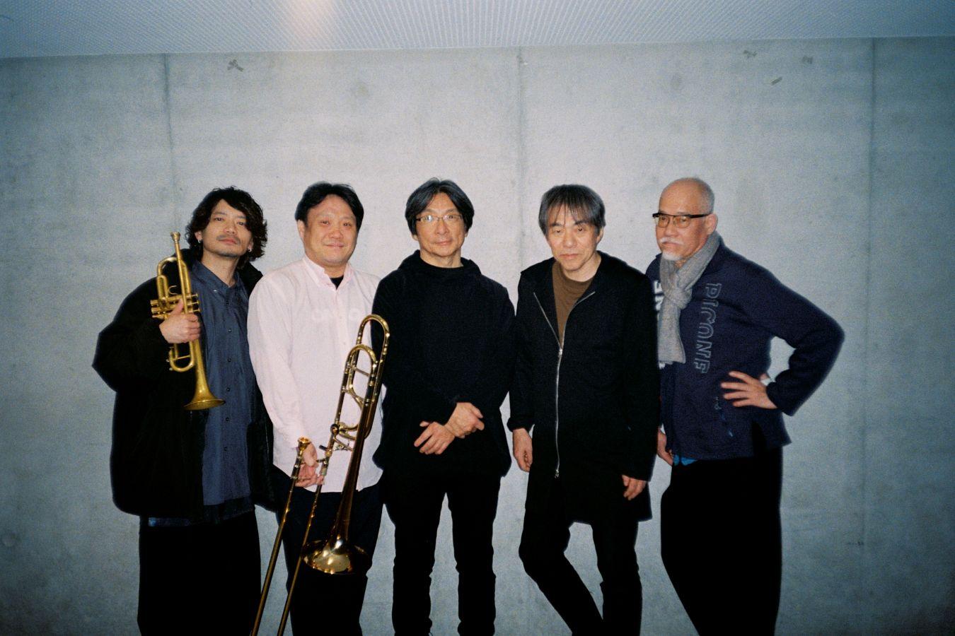 Five people stand in front of a gray wall; two hold instruments, a trumpet and a trombone.
