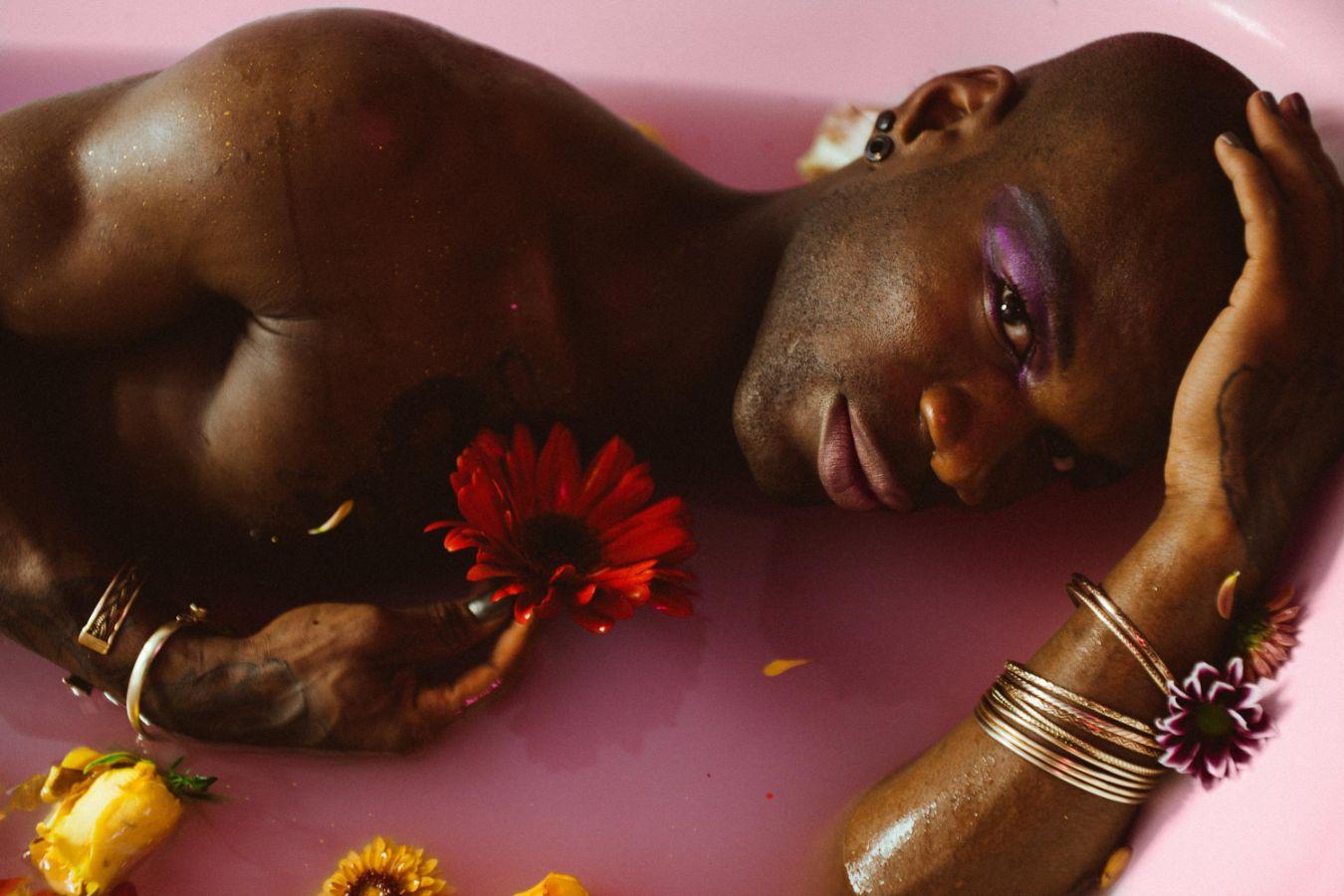 Foto von einer Person, die in einer rosafarbenen Badewanne mit rosafarbenem Wasser und Blumen liegt.